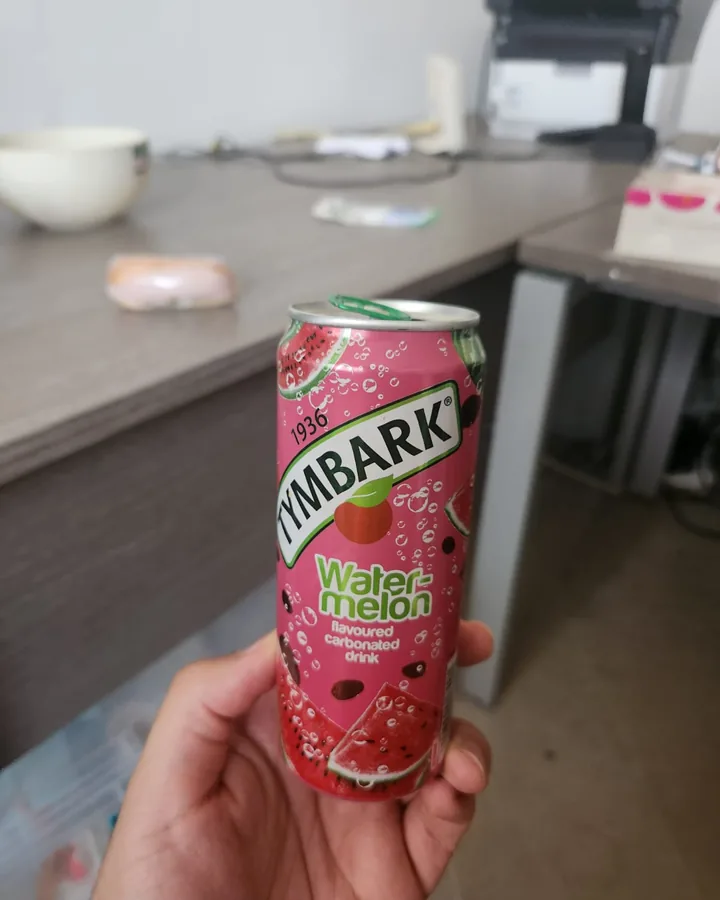 A hand holding a watermelon drink can in a cluttered office